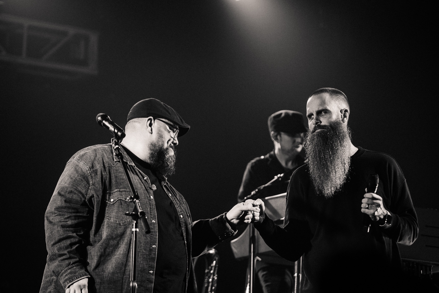 Two foreground speakers sharing a fist bump onstage, with a musician behind them